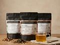 Three black tea bags with labels on a wooden surface next to a cup of tea.
